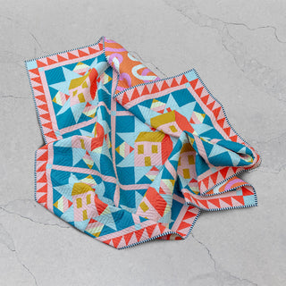 Brightly colored quilt with geometric house and triangle designs folded on a cracked concrete surface, showcasing the Cottagecore Quilt Pattern (Download) by Toad & Sew.