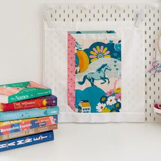 Colorful fabric quilt block featuring a horse and floral design displayed on a pegboard, with a stack of books nearby, showcasing the Currently Reading Block Pattern (Download) by Lou Orth Designs.