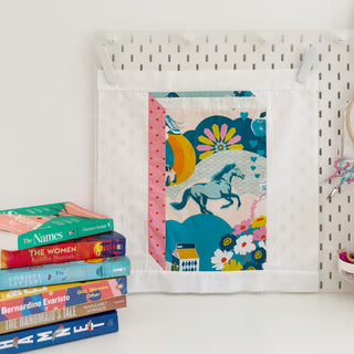 Quilt block with colorful horse and floral design hanging on pegboard, styled next to a stack of books, showcasing The Book Block Collection — Complete 5-Block Bundle (Download) by Lou Orth Designs.