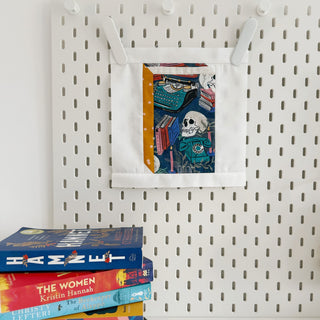Mini quilt block featuring a colorful vintage skull and typewriter design, hung on a pegboard with a stack of books nearby, showcasing Currently Reading Block Pattern (Download) by Lou Orth Designs.