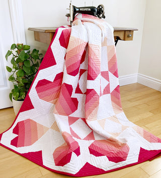 Quilt draped over a vintage sewing machine featuring a geometric pink and red heart design, showcasing the Dancing Hearts Quilt Pattern (Download) by Penny Spool Quilts.