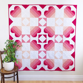 Bright quilt featuring gradient pink hearts and geometric blocks hanging on a wall with a plant nearby, showcasing Penny Spool Quilts Dancing Hearts Quilt Pattern (Download).