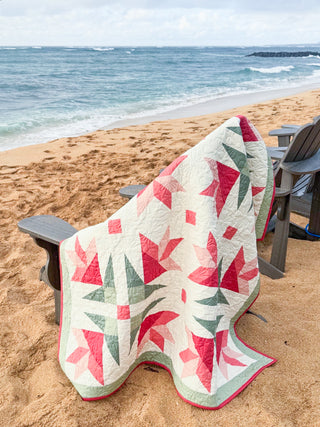 Colorful floral quilt draped over a beach chair on sandy shore, showcasing Dancing Lilies Quilt Pattern (Download) by Shadow Valley Quilts in soft pink, red, and green tones.