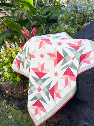 Colorful geometric quilt with pink, coral, and green lilies pattern, draped over a rock in a garden, showcasing the Dancing Lilies Quilt Pattern (Download) by Shadow Valley Quilts.