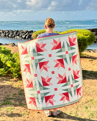 Person holding a colorful quilt featuring pink lilies and green geometric accents outdoors by the ocean, showcasing the Dancing Lilies Quilt Pattern (Download) by Shadow Valley Quilts.