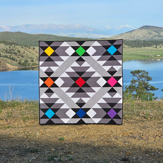 Desert Chill Quilt Pattern by Staci W Quilts cover: dark grey quilt with multicolored diamond blocks in yellow, green, blue, orange, and red, displayed by a mountain lake.