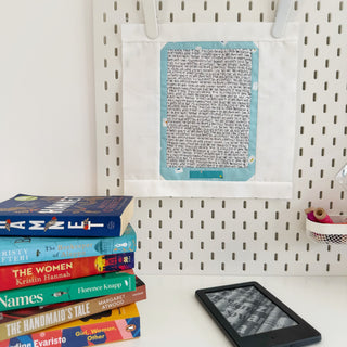 Stack of colorful books next to an E Reader with a fabric quilt block titled E Reader Block Pattern (Download) by Lou Orth Designs hanging on a pegboard wall.