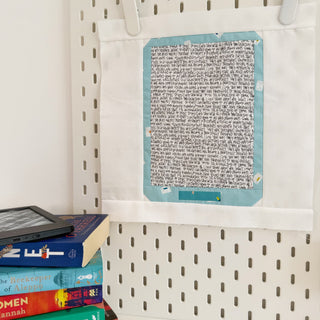 Fabric quilt block featuring handwritten text design with a turquoise border hanging on a pegboard, paired with a stack of colorful books and an e-reader, showcasing The Book Block Collection — Complete 5-Block Bundle (Download) by Lou Orth Designs.