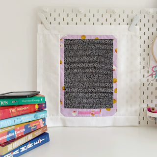 Wall hanging quilt block with purple border featuring small fruit motifs, displayed on a pegboard with a stack of colorful books nearby, showcasing The Book Block Collection — Complete 5-Block Bundle (Download) by Lou Orth Designs.