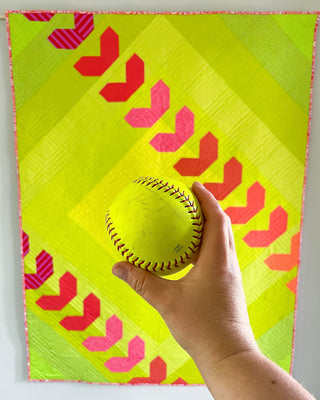 Fastball 2.0 Quilt Pattern (Download) by Jessica Steele Design showing a neon green baseball quilt with pink and red chevrons and a hand holding a softball