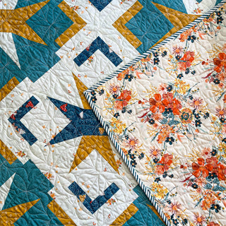 Close-up of a colorful quilt with geometric star and block patterns in blue, mustard, and white, paired with a floral backing, showcasing the Festive Gala Quilt Pattern (Download) by A Piece of Quiet Quilts.