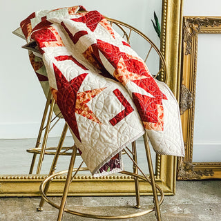 Red and cream geometric quilt draped over a gold metal chair beside an ornate gold frame, showcasing the Festive Gala Quilt Pattern (Download) by A Piece of Quiet Quilts.