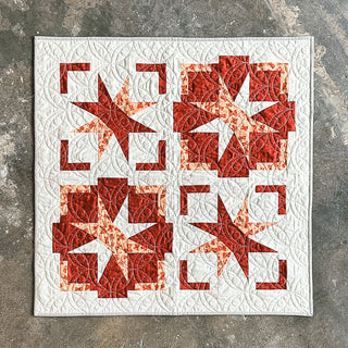 Festive Gala Quilt Pattern by A Piece of Quiet Quilts cover: red and white geometric star quilt shown flat on a textured stone surface.
