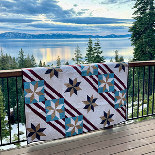 Fifty Nifty Quilt Pattern (Download) by Shadow Valley Quilts, featuring a geometric star and stripe design in blues, browns, and burgundy, displayed overlooking a serene lake view.