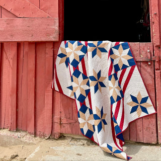 A patriotic star and stripe quilt draped over a rustic red barn door, showcasing the Fifty Nifty Quilt Pattern (Download) by Shadow Valley Quilts in red, blue, tan, and white colors.