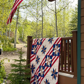 A red, white, and blue star and stripe quilt made with the Fifty Nifty Quilt Pattern (Download) by Shadow Valley Quilts draped over a porch railing with green trees and an American flag in the background.