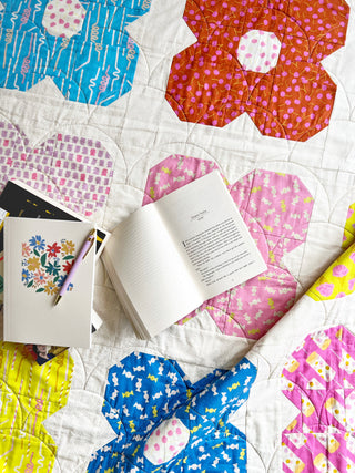 Close-up of a colorful quilt with floral and geometric patterns in pink, blue, yellow, and orange fabric squares, featuring an open book and a notebook, designed by Lou Orth Designs using the Flower Friends Quilt Pattern (Download).
