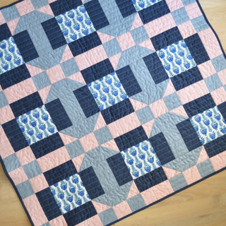 Close-up of a colorful quilt featuring geometric shapes in pink, blue, and gray with floral fabric squares, made using the Footpath Quilt Pattern (Download) by Sewn.