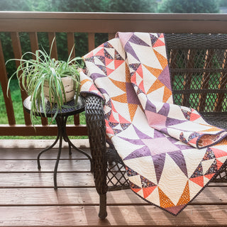 A colorful geometric quilt draped over a wicker chair on a wooden porch next to a small table with a potted plant, showcasing the Freya June Quilt Pattern (Download) by A Piece of Quiet Quilts.