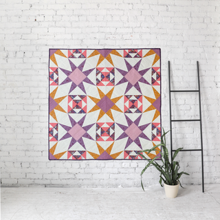 Freya June Quilt Pattern by A Piece of Quiet Quilts cover: purple, pink, and orange geometric star quilt hanging on a white brick wall with a metal ladder and plant.