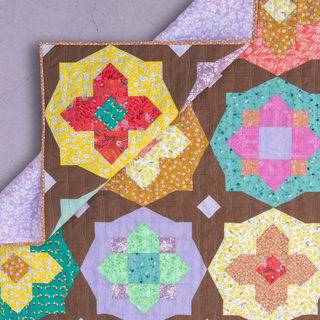 Close-up of a colorful quilt with floral and geometric patterns in bright tones, featuring a folded corner showing a lilac floral backing, illustrating the Garden Glow Quilt Pattern (Download) by Toad & Sew.