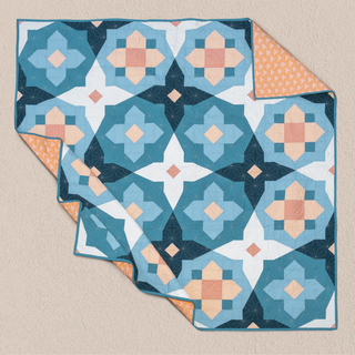 Folded quilt featuring geometric floral and cross patterns in soft blue, peach, and navy tones with a contrasting orange triangular print backing, designed by Toad & Sew, showcasing the Garden Glow Quilt Pattern (Download).