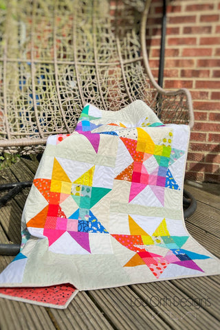 Colorful star quilt with bright fabric patterns draped over a wicker chair outdoors, showcasing the Gemburst Quilt Pattern (Download) by Lou Orth Designs.