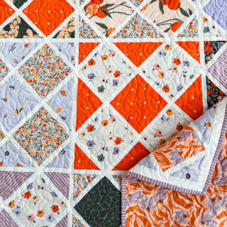 Close-up of a colorful quilt featuring orange, purple, and floral fabric squares in a geometric layout, showcasing the Harlequin Bloom Quilt Pattern (Download) by A Piece of Quiet Quilts.