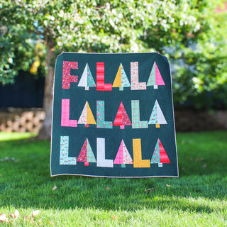 Fa-La-La Quilt Pattern (Download) colorful holiday quilt with repeated geometric lettering displayed outdoors by Patternly Collective