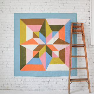 Modern geometric quilt hanging on a white brick wall with a wooden ladder nearby, showcasing the Arete 8-Color Quilt Pattern (Download) by Eudaimonia Studio.