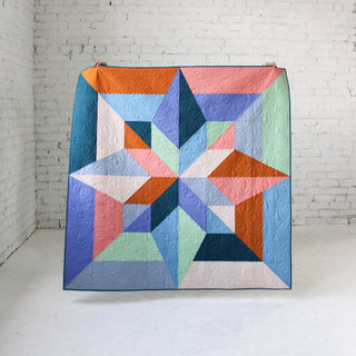 Colorful geometric quilt displayed against a white brick wall, showcasing detailed stitching and a modern design made with the Arete 8-Color Quilt Pattern (Download) by Eudaimonia Studio.