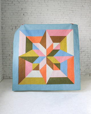 Colorful geometric quilt with star and diamond shapes in pastel and earth tones, displayed against a white brick wall, featuring Arete 8-Color Quilt Pattern (Download) by Eudaimonia Studio.