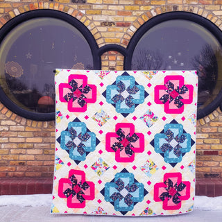 Colorful quilt with bright pink and blue geometric floral blocks and a white background, displayed outdoors, illustrating the Ixora Quilt Pattern (Download) by Thai Charm.
