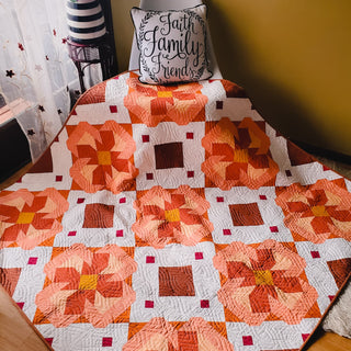 A vibrant orange and white quilt with geometric floral blocks draped over a chair, displaying the Ixora Quilt Pattern (Download) by Thai Charm, styled in a cozy room.