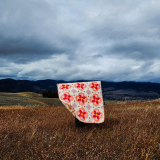 Person standing in tall grass holding a pink and red floral quilt with geometric blocks against a moody cloudy sky, showcasing the Ixora Quilt Pattern (Download) by Thai Charm.