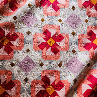 Close-up of a colorful quilt with mauve, coral, red, and mustard geometric shapes and intricate stitching featuring the Ixora Quilt Pattern (Download) by Thai Charm.