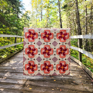 Colorful floral quilt with geometric shapes in shades of mauve, orange, and red displayed outdoors on a rustic wooden bridge, showcasing the Ixora Quilt Pattern (Download) by Thai Charm.