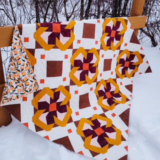 Colorful yellow, purple, and brown floral patterned quilt displayed outdoors on a wooden fence in snow, showcasing the Ixora Quilt Pattern (Download) by Thai Charm.