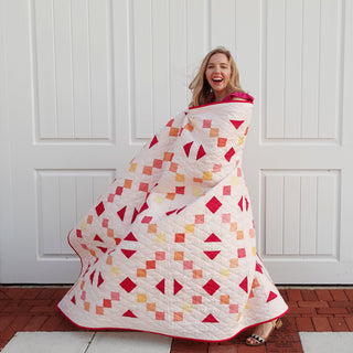 Keystone Quilt Pattern (Download) by Homemade Emily Jane featuring a modern geometric quilt with balanced symmetry and colorful red peach and gold shapes wrapped around a woman