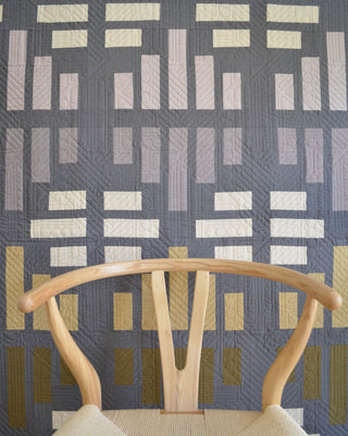 Modern geometric quilt with rectangular blocks in gray, beige, and mustard tones showcased behind a light wood chair, featuring the Leaded Light Quilt Pattern (Download) by Sewn.