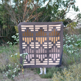 Person standing outdoors holding a geometric quilt made with earthy tones, showcasing the Leaded Light Quilt Pattern (Download) by Sewn in a natural setting.
