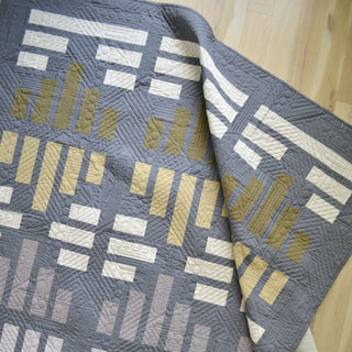 Modern quilt featuring geometric blocks in muted gold, cream, and gray tones with detailed quilting patterns, showcasing Sewn's Leaded Light Quilt Pattern (Download).