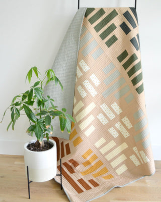 Leaded Light Quilt Pattern (Download) by Sewn displayed on a modern quilt rack with neutral tones and geometric block design, next to a green leafy plant.