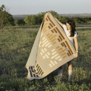 Young woman outdoors holding a neutral-toned quilt with geometric patterns, showcasing the Leaded Light Quilt Pattern (Download) by Sewn in a natural setting.