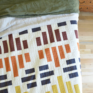 Close-up of a handmade quilt with a geometric pattern in brown, orange, navy, and yellow on a cream background, styled casually on a wooden floor, showcasing the Leaded Light Quilt Pattern (Download) by Sewn.