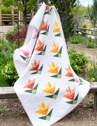 White quilt with colorful geometric lily block patterns in pink, orange, and yellow tones, draped outdoors on a wooden bench, showcasing Lilies on Parade Quilt Pattern (Download) by Shadow Valley Quilts.