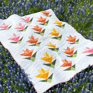 Quilt featuring colorful lilies in orange, yellow, and pink on a white background, laying in a field of blue flowers, showcasing Lilies on Parade Quilt Pattern (Download) by Shadow Valley Quilts.