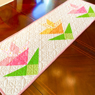Table runner featuring a quilted floral design with pink and yellow lilies and green geometric leaves, showcasing Shadow Valley Quilts' Lilies on Parade Quilt Pattern (Download).