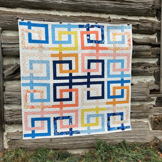 Colorful modern quilt with interlocking Greek meander-inspired ribbons in blue, yellow, orange, and white, showcasing the Line Dance Quilt Pattern (Download) by Penny Spool Quilts.