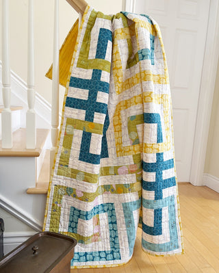 Quilt hanging over a stair railing featuring geometric blue, green, and yellow ribbons in a modern greek meander style by Penny Spool Quilts, showcasing the Line Dance Quilt Pattern (Download).